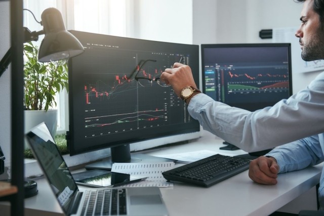 Young trader is pointing at graphs on computer screen and analyzing data while working in his modern office
